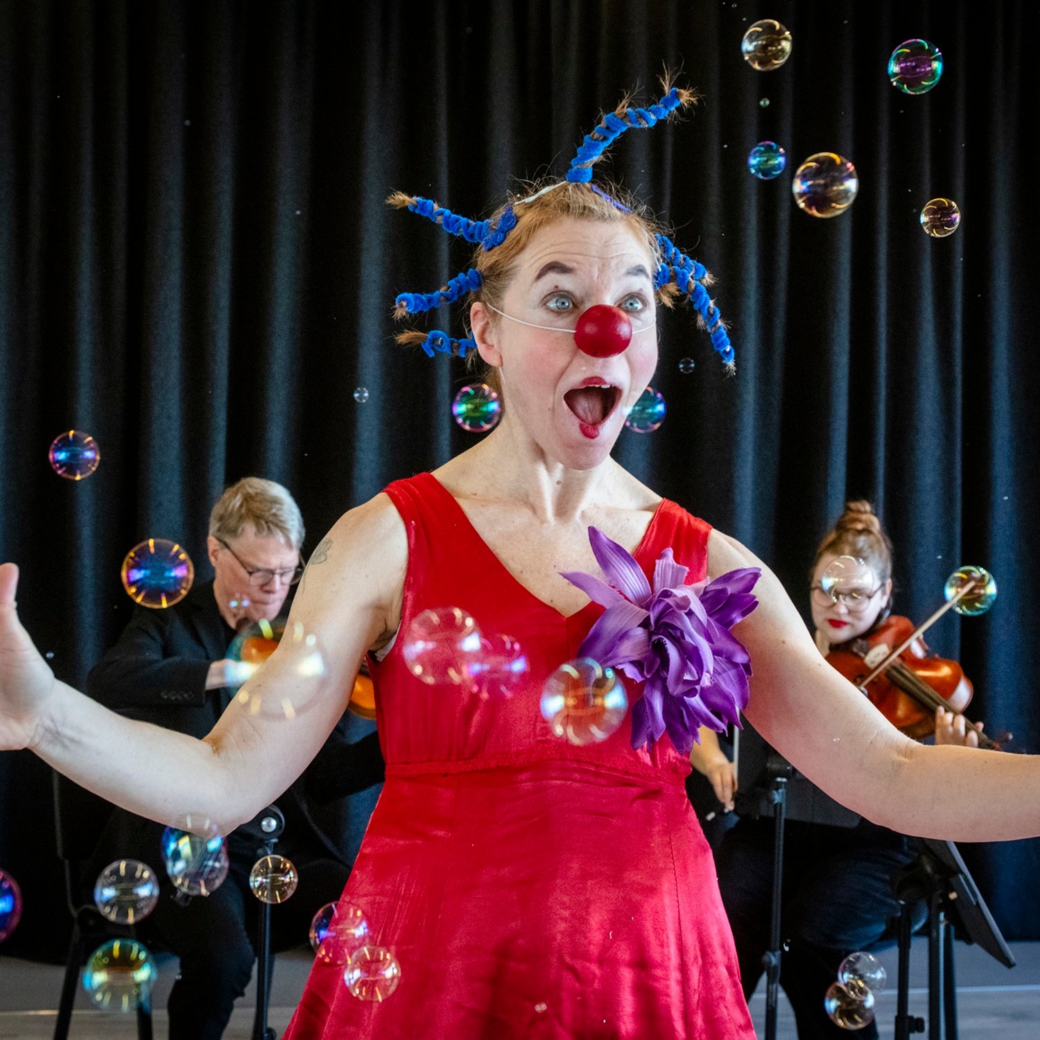 Framför en stråkorkester står en clown med röd näsa.