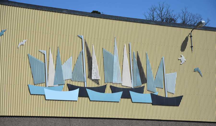 Konstverk bestående av flera segelbåtar i olika nyanser av vit, grått och blått mot en gul fabriksvägg. Båtarna är gjorda i metall och fastsatta på väggen. 