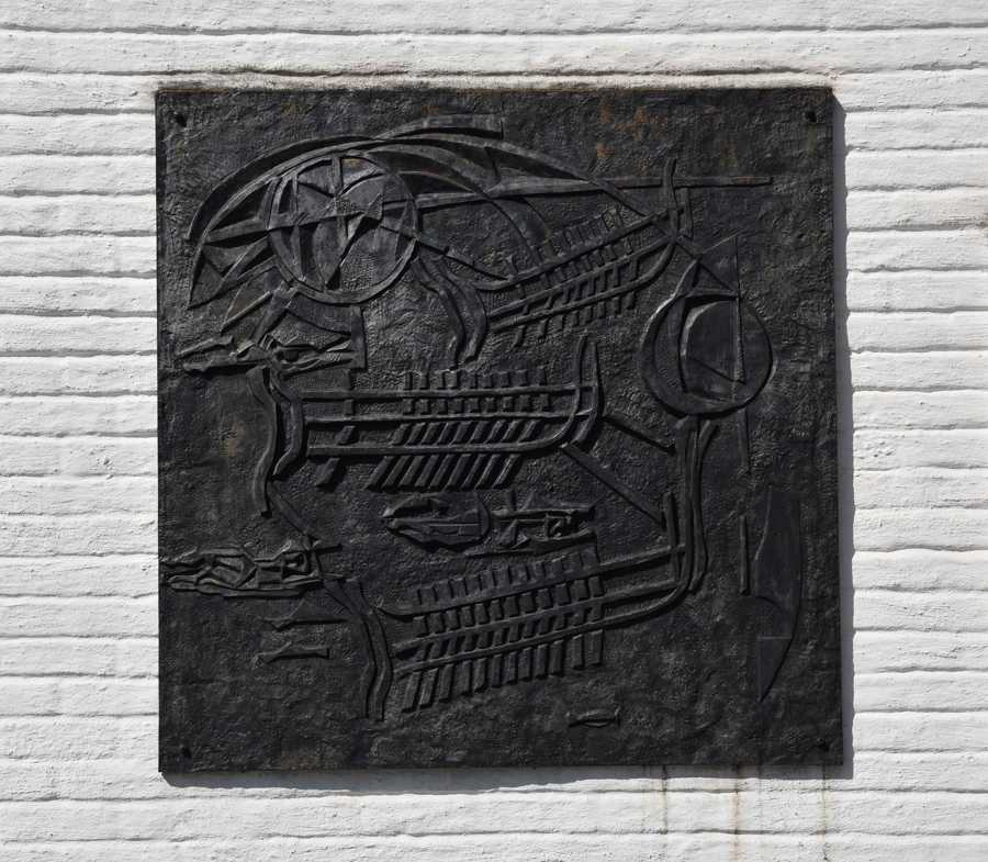 Relief i järn med olika figurer och symboler. Är monterad mot en vit tegelvägg. 