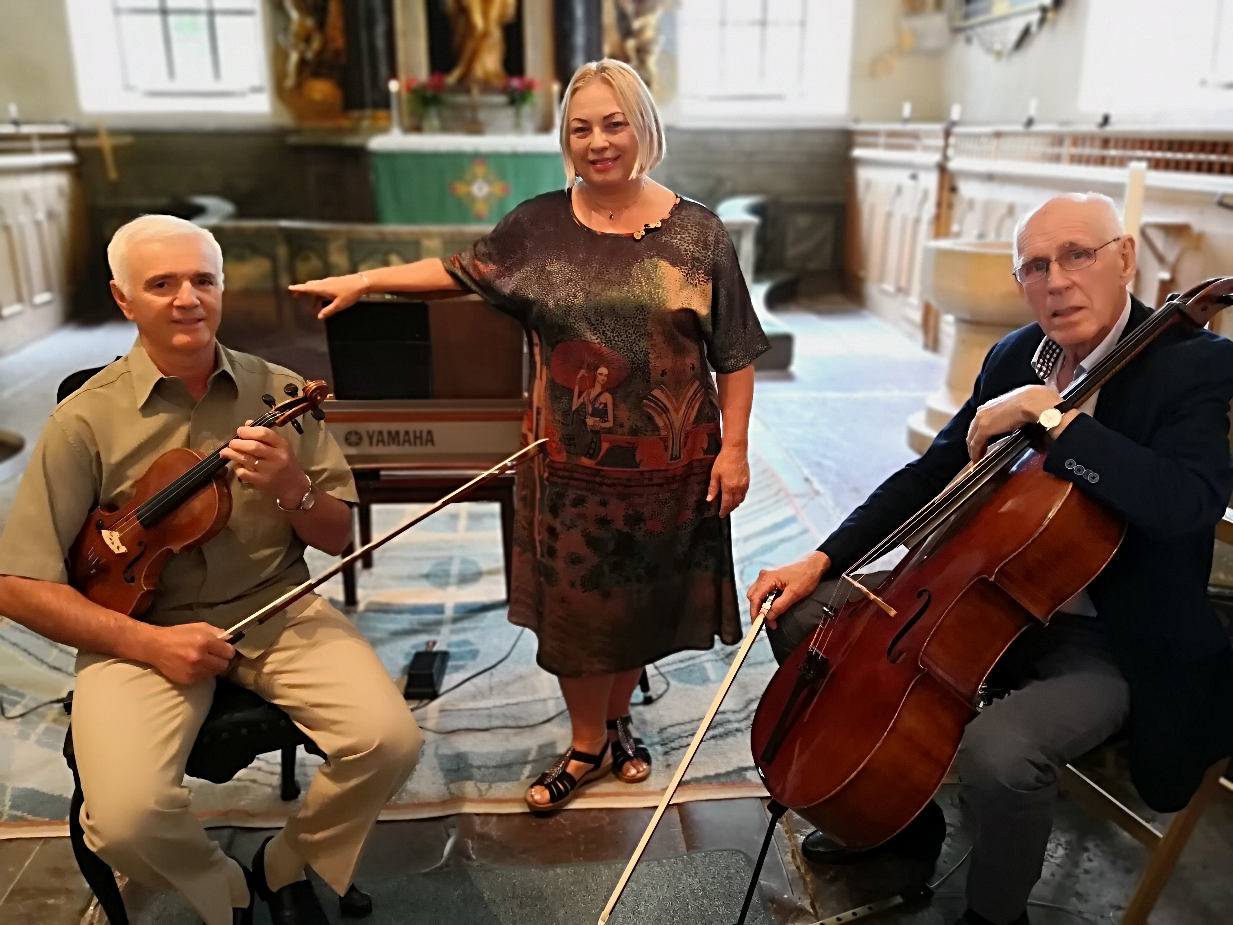 Ensembeln visar upp sig i en kyrka. Det är 3 personer i bilden som är vid sina instrument, fiol, piano, cello