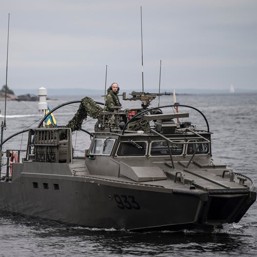 pressbild från Försvarmakten på en av marinens båtar med en soldat som tittar in i kameran och flera soldater i förarhytten